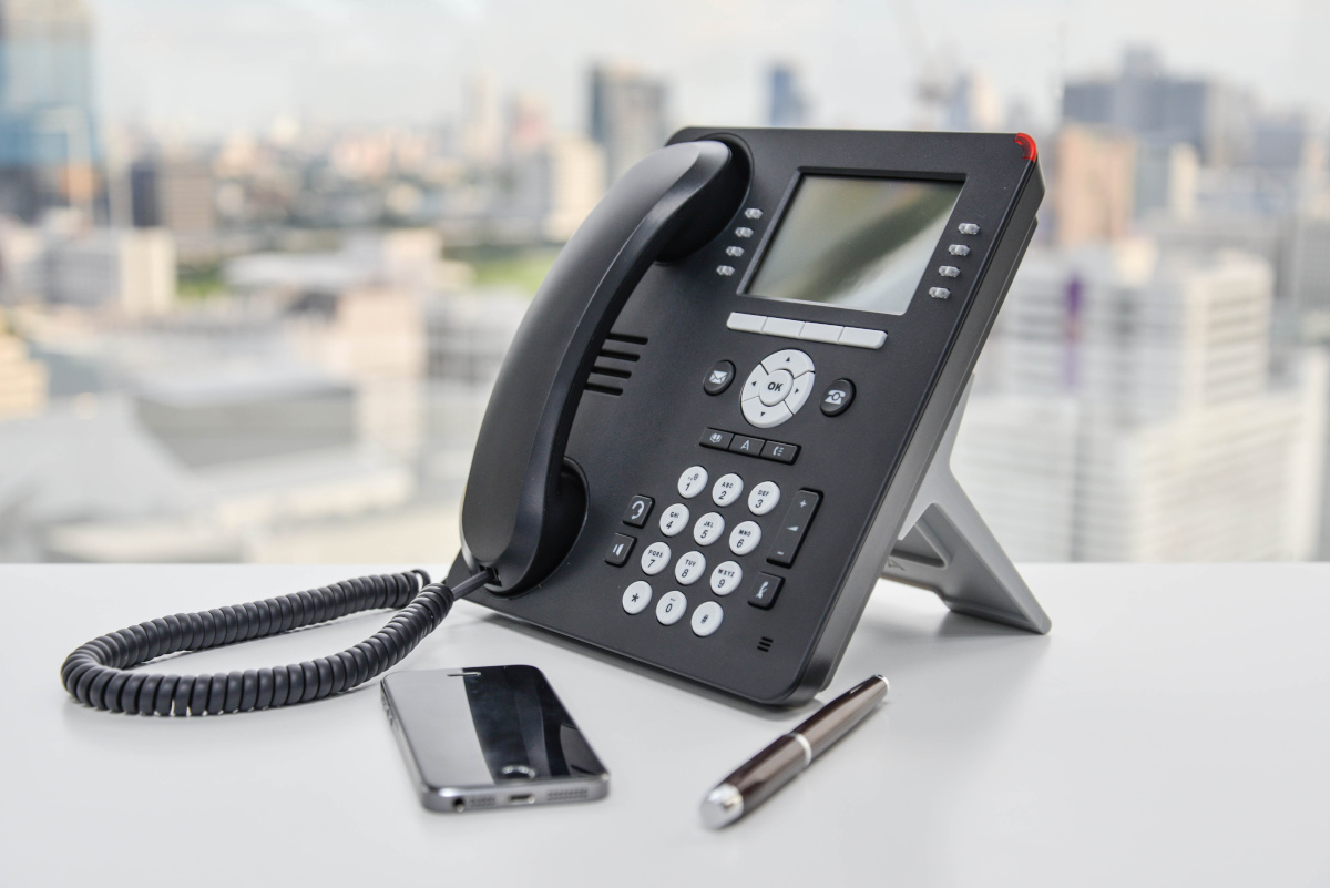 A modern business phone on a desk next to a cell phone and a pen