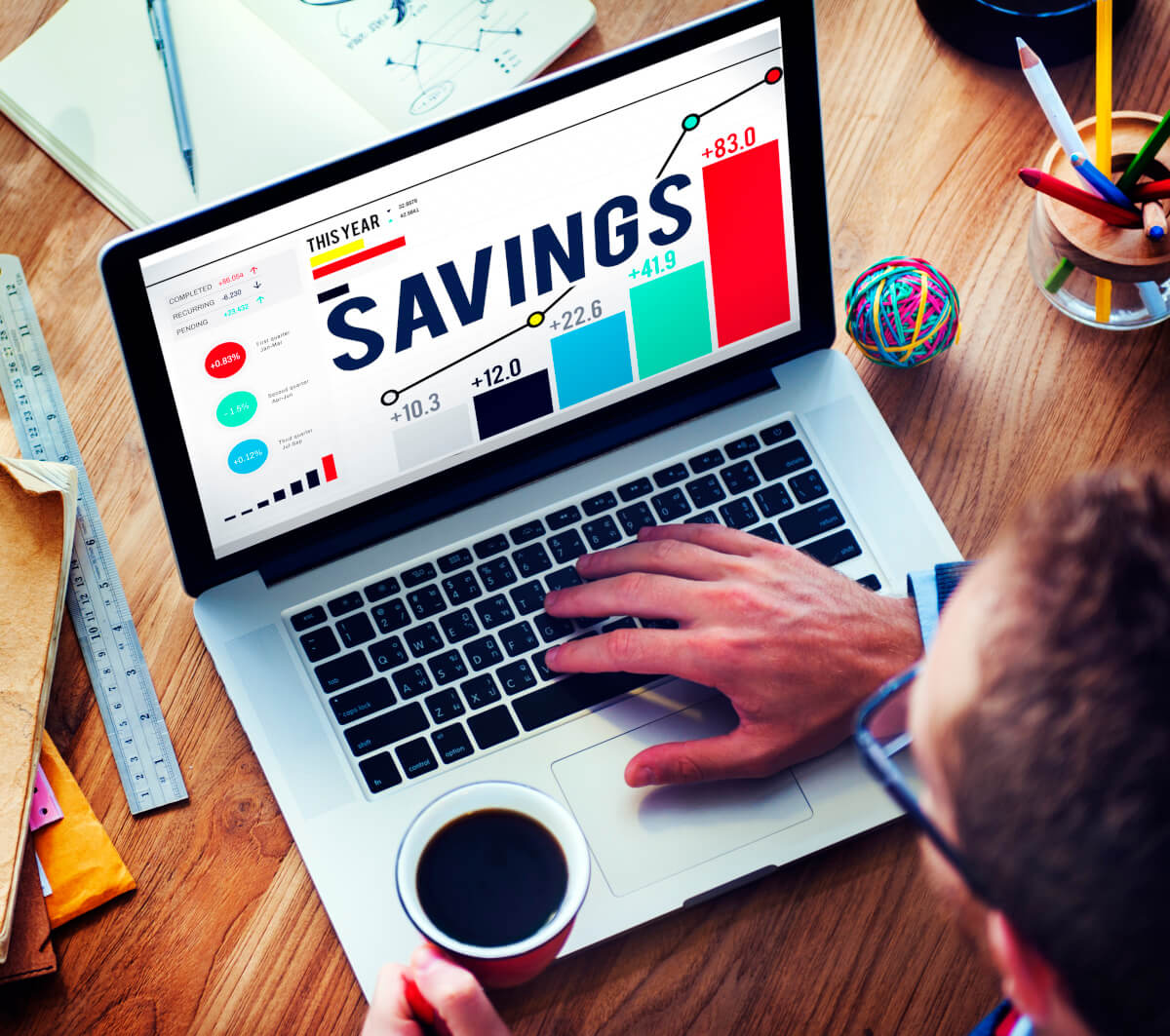 A man in glasses working on a laptop that says savings, while displaying info through graphs, while also holding a coffee in his other hand