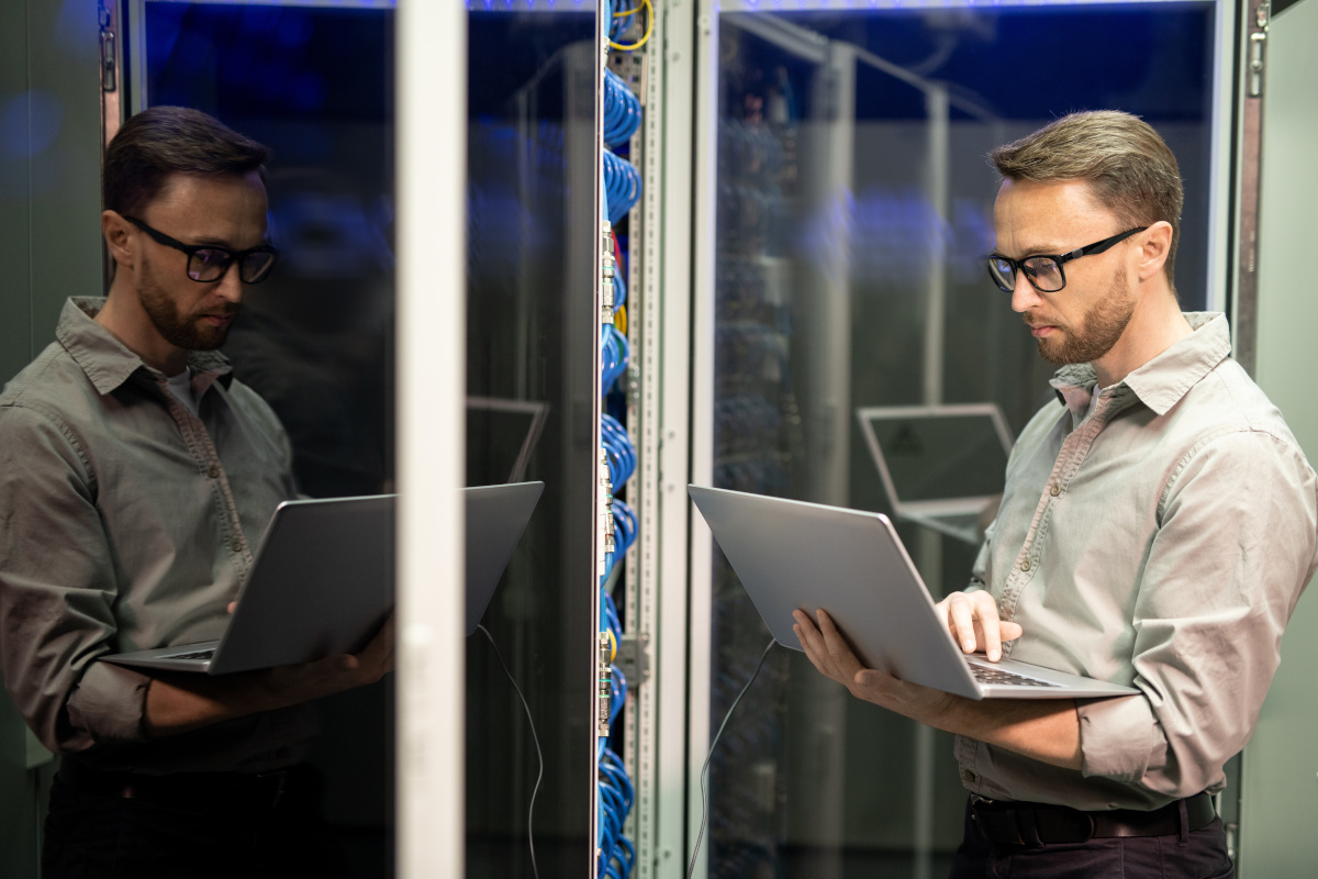 Serious busy young IT engineer with beard standing by server cabinet and using laptop while providing network support.